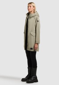 Žena stojící před jednobarevným pozadím, oblečená v béžovém dlouhém kabátu s kapucí a klopovými kapsami, černých legínách a černých šněrovacích bojových botách.