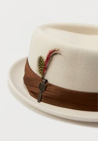 Krémový plstený klobúk fedora s hnedým pásikom, ozdobený žltými škvrnami a červenými perami, ktoré sú upevnené kovovou ihlou na bielom pozadí.