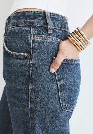 Ręka w tylnej kieszeni niebieskich dżinsów, z wieloma złotymi i srebrnymi bransoletkami na nadgarstku, na białym tle.