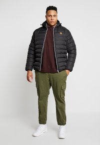 Sort pufferjakke med lynlås, sølvdetaljer og hætte; parret med en maroon sweatshirt og oliven cargo-bukser. Hvide sneakers fuldender outfittet.