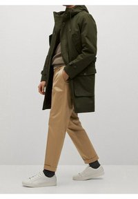 Olivengroene parka met een capuchon, voorzien van zakken en knoopaccenten. Gecombineerd met lichtbruine broeken en witte sneakers.