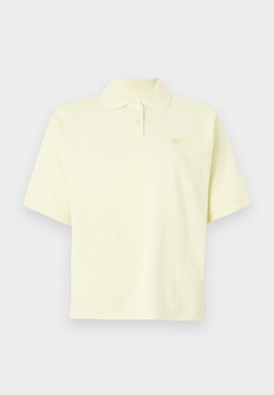 Lühikeste varrukatega heledast kollasest polokuuest, millel on kaks nuppu ja väike südamemotiiv vasakul rinnal, tavalise tausta ees.