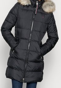 Černý prošívaný zimní kabát s kapucí lemovanou kožešinou, předním zipem, bočními kapsami a malým logem na hrudi, nošený přes modré džíny.