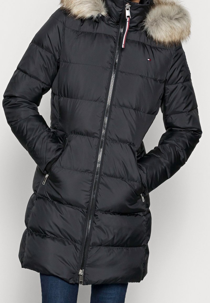 Černý prošívaný zimní kabát s kapucí lemovanou kožešinou, předním zipem, bočními kapsami a malým logem na hrudi, nošený přes modré džíny.