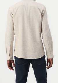 Camisa de manga larga color beige claro con cuello abotonado, textura sutil y puños de un solo botón, combinada con jeans azul oscuro.