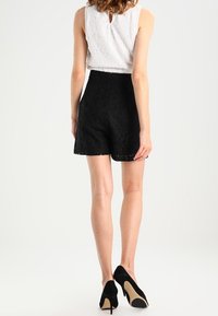 Černé krajkové šortky s vysokým pasem, s vroubkovaným lemem a strukturovaným vzorem. Kombinovány s černými vysokými podpatky a bílým topem.