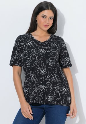 Frau mit langem dunklem Haar, die ein schwarzes kurzärmeliges Shirt mit weißen abstrakten Linien- und Figurenmustern trägt, kombiniert mit blauen Jeans.