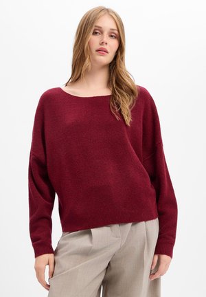 Jeune femme portant un pull bordeaux ample et un pantalon beige à jambes larges, debout devant un fond blanc.