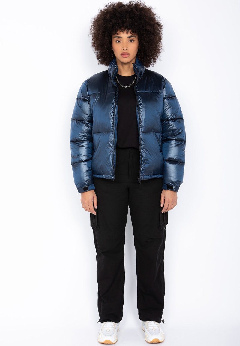 Doudoune bleu marine avec texture brillante, col montant et fermeture éclair. Portée par-dessus un haut noir, associée à un pantalon cargo noir et des baskets blanches.