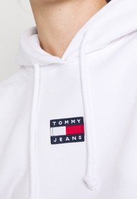 Sudadera blanca con capucha que presenta un logo de parche de tela con acentos en rojo, blanco y azul que dice "TOMMY JEANS". Textura suave con cordones.