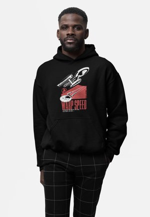 Man draagt een zwarte hoodie met een rood-witte Star Trek warp speed afbeelding en een zwarte broek met een wit rasterschema.