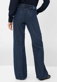 Zadní pohled na osobu v tmavě modrých širokých džínách a černých síťovaných botech, s jednou rukou opřenou o bok na bílém pozadí.