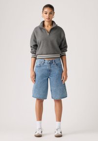 Γυναίκα που φοράει γκρι φούτερ με φερμουάρ μέχρι το στήθος και ριγέ μανσέτες, σορτς τζιν μέχρι το γόνατο, λευκές κάλτσες και καθημερινά αθλητικά παπούτσια, στέκεται μπροστά σε λευκό φόντο.