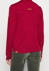 Chemise rouge à manches longues avec un motif texturé, dotée d'un col rond, d'accents dorés et d'une étiquette de logo en bas. Portée avec un pantalon vert olive.