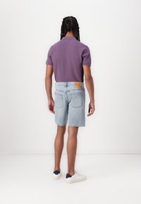 Polo shirt viola, pantaloni corti in denim azzurro chiaro con etichetta sul retro e sneakers bianche con dettagli navy. Vista posteriore, sfondo neutro.