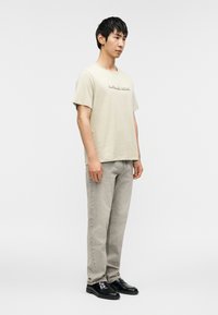 Ανοιχτό μπεζ βαμβακερό μπλουζάκι με ανάγλυφο λογότυπο, συνδυασμένο με γκρι τζιν παντελόνι και μαύρα παπούτσια. Το μοντέλο στέκεται σε προφίλ μπροστά σε απλό φόντο.