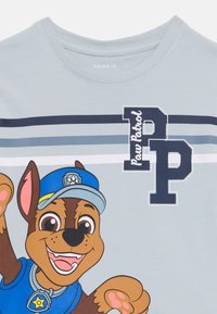 Šviesiai mėlynas marškinėlis su dideliu animaciniu šunimi mėlynu uniforma. Tekstas skelbia „Paw Patrol“ su tamsiai mėlynomis juostomis per dizainą.