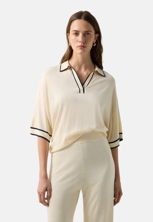 Vrouw met stijlbruin haar draagt een crèmekleurige gebreide top met zwarte bies op de kraag en mouwen, gecombineerd met bijpassende gebreide broek.