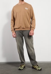 Sudadera marrón con texto "Fresh Coffee", combinada con vaqueros gris deslavado y zapatillas de camuflaje verde. Textura suave con puños acanalados.