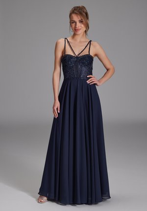 Femme en robe de soirée bustier bleu marine ornée de perles, avec une jupe fluide, debout, une main sur la hanche, sur fond gris.