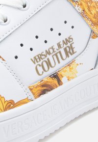 Λευκό αθλητικό παπούτσι με χρυσές λεπτομέρειες και ανθισμένο σχέδιο. Διαθέτει επενδυμένη γλώσσα, διάτρητο κουτί δαχτύλων και υφή με το λογότυπο στη πλευρά.