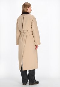 Cămașă lungă bej cu talie curea, având un guler maro închis, design cu spate despicat și țesătură ușor texturată. Purtați cu pantaloni negri și cizme.