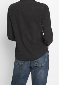 Black denim shirt with a collar and long sleeves, featuring a curved hem and back yoke detail. Paired with blue jeans.