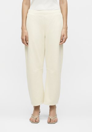 Γυναίκα που φοράει φαρδιά κρεμ παντελόνια και μπεζ πέδιλα με τετράγωνη μύτη και τακούνι, στέκεται μπροστά σε απλό φόντο.