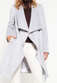 Cappotto in misto lana grigio chiaro con davanti aperto, colletto drappeggiato, maniche lunghe e due tasche laterali, indossato sopra una maglia marrone e pantaloni bianchi.