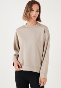 Felpa beige a maniche lunghe con colluccio rotondo, vestibilità comoda e orlo a coste. Texture liscia e design minimal, indossata con jeans scuri.
