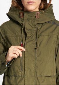 Olivgrön, dragkedjeförsedd jacka med hög krage och justerbara snoddar. Materialet verkar ha en texturerad yta med synliga sömnadsdetaljer.