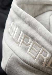 Grå sweatshirtstof med en foldet krave og det broderede ord "SUPER" i hvid. Baggrunden består af et mørkere tekstil.