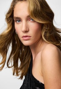 Modelka o długich, falujących blond włosach, ubrana w czarny, cekinowy top na jedno ramię, prezentujący błyszczącą fakturę i szczegółowe akcenty.