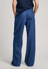 Osoba oblečená v modrých džínách s vysokým pasem a širokými nohavicemi a námořnickém pleteném topu, stojící s rukama v kapsách, otočená zády.