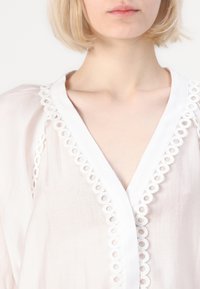 Blusa in tessuto leggero e trasparente di colore chiaro con scollatura a V e delicati bordi di pizzo circolare bianco lungo il collo e le maniche.