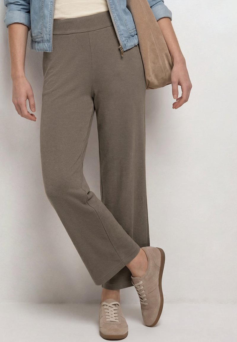 Persona con pantalones anchos taupe, zapatillas beige, chaqueta vaquera azul claro, llevando bolso tote de ante beige, de pie frente a una pared blanca.
