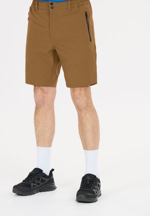 Braune Herren-Freizeithosen mit vorderer Reißverschlusstasche, kombiniert mit weißen Socken und schwarzen Sportschuhen vor einfachem Hintergrund.