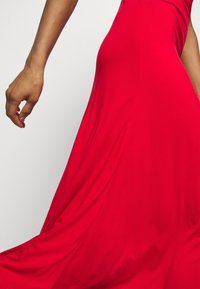 Czerwona sukienka o średniej długości z dopasowanym topem i swobodnym dołem, wykonana z gładkiego materiału. Ręka trzyma materiał, podkreślając jego ruch.
