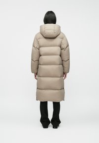 Beige puffer coat with a hood, featuring a quilted design, long silhouette, and a smooth texture. Worn over black wide-leg trousers.