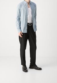 Mies, joka on pukeutunut vaaleansiniseen nappipaitaan valkoisen toppatakin päällä, mustiin housuihin vyöllä ja mustiin juhlakengiin, seisoo valkoista taustaa vasten.