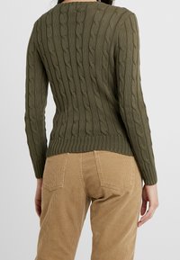 Olivgrüner, gestrickter Pullover mit Zopfmuster, gerippten Bündchen und Saum. Kombiniert mit beigen Cordhosen, die Gesäßtaschen zeigen.