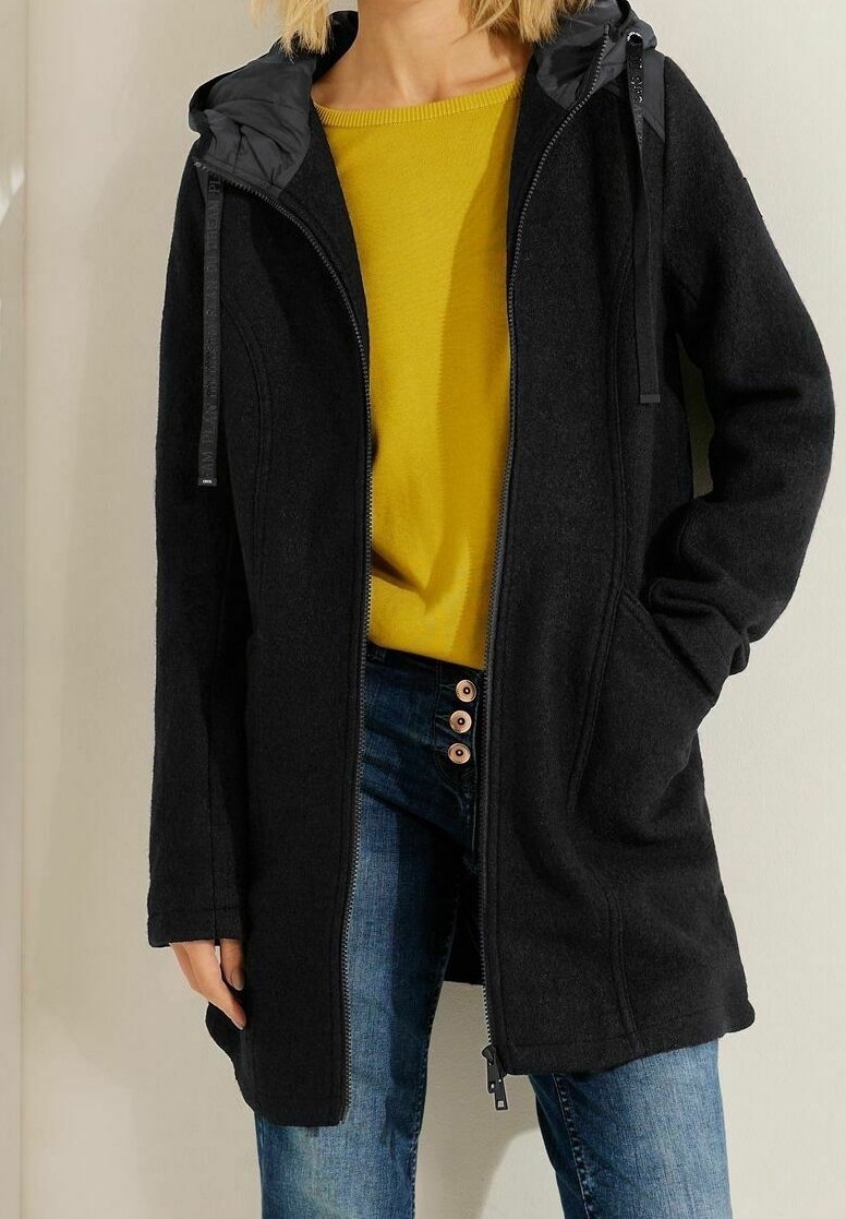 Chaqueta larga negra con capucha desmontable, con cremallera frontal y bolsillos laterales. Llevada sobre una camiseta amarilla y vaqueros azules, tejido texturizado.