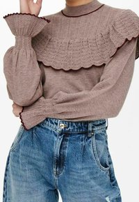 Pull en maille marron avec des détails de volants texturés au niveau du col et des poignets, associé à un jean en denim bleu déchiré taille haute.