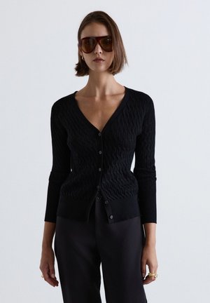 Donna con occhiali da sole marroni oversize, cardigan nero testurizzato con bottoni e pantaloni scuri, in piedi davanti a uno sfondo chiaro e uniforme.