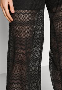 Gros plan sur une main reposant sur un tissu en dentelle noire transparente avec des motifs en zigzag et floraux sur une peau nue.