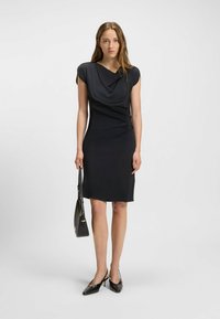 Schwarzes Kleid mit drapiertem Ausschnitt und kurzen Ärmeln. Figurbetonte Silhouette, gerafftes Detail an der Seite, mit glatter Textur. Getragen mit schwarzen High Heels und Handtasche.