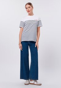 T-shirt blanc et navy à manches courtes rayé, associé à un jean en denim bleu à larges jambes et des sneakers gris avec accents blancs. Marque simple visible.