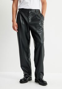 Pantaloni in finta pelle nera con una vestibilità rilassata, caratterizzati da una piega frontale, tasche laterali e una finitura liscia e lucida. Completati da stivali neri.