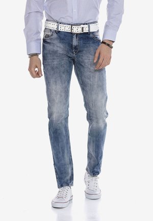 Jean bleu délavé à coupe slim, avec une ceinture cloutée blanche, un design classique à cinq poches, et une texture usée, associé à des baskets blanches.