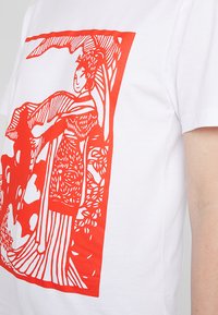 Vit bomull t-shirt med en djärv röd grafisk tryck av en figur omgiven av blommiga och linjära mönster. Korta ärmar och rund halsringning.